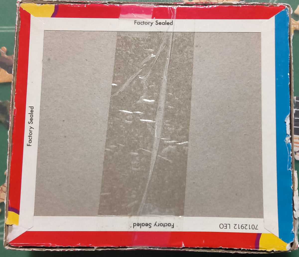 A square cardboard box insert or packaging panel with a gray reflective center and red, white, and blue borders, placed on a green jigsaw puzzle mat with scattered puzzle pieces around it.