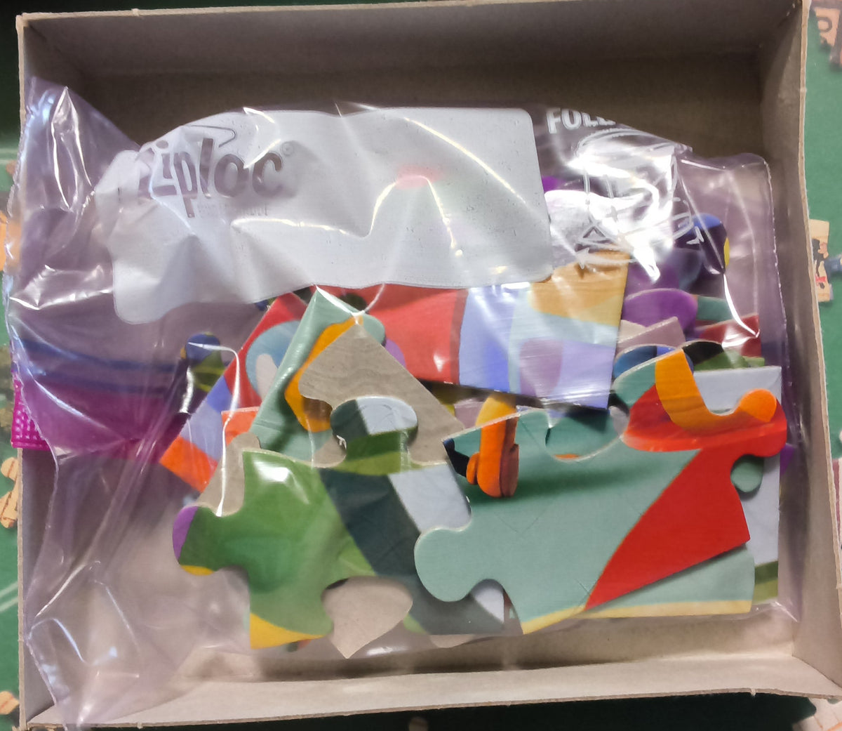 A box of colorful jigsaw puzzle pieces inside a clear plastic bag, shown on a green puzzle mat with scattered puzzle pieces around it and a puzzle box partially visible on the right.