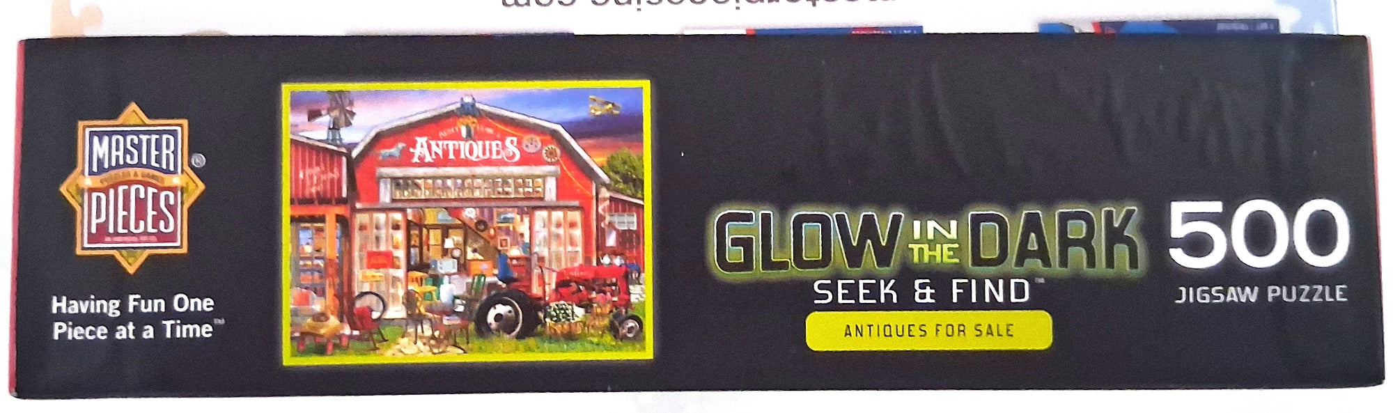 A 500-piece jigsaw puzzle box by Master Pieces featuring a red barn and tractor illustration, with text reading 'Glow in the Dark Seek & Find' and 'Antiques for Sale.'.