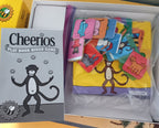 Cheerios Play Book Bingo Game set shown in its box, featuring a gray game book with a cartoon monkey and a clear zippered bag filled with colorful bingo cards.