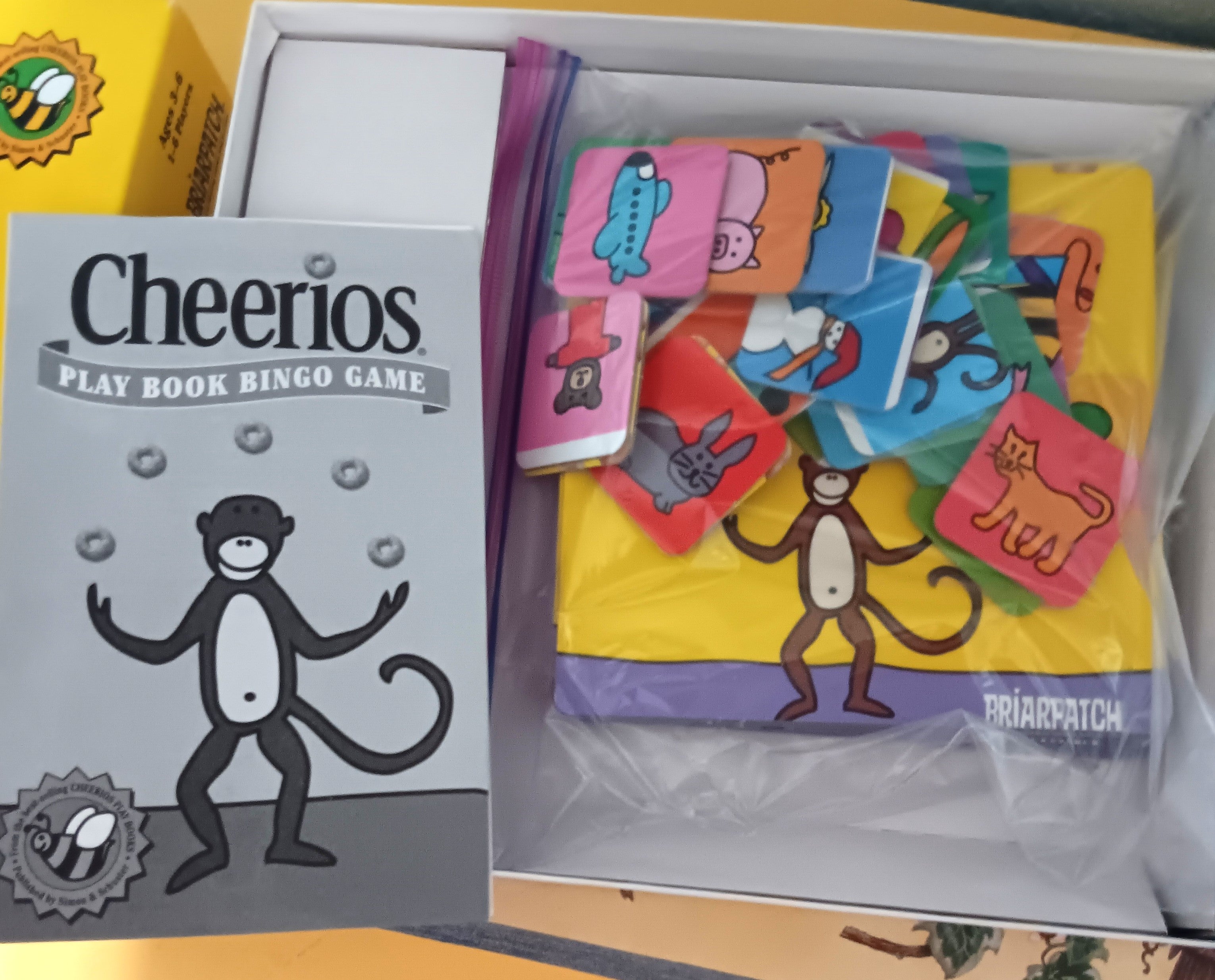 Cheerios Play Book Bingo Game set shown in its box, featuring a gray game book with a cartoon monkey and a clear zippered bag filled with colorful bingo cards.
