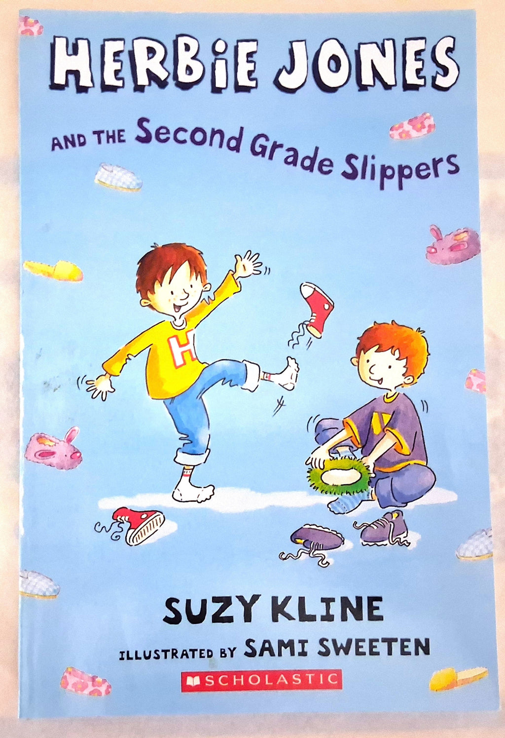 Front cover of the children's book 'Herbie Jones and the Second Grade Slippers' by Suzy Kline, illustrated by Sami Sweeten, published by Scholastic. The cover features a light blue background with cartoon illustrations of two children in colorful clothing and several pairs of mismatched slippers.
