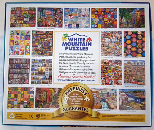 The back cover of a White Mountain Puzzles jigsaw puzzle box, featuring a grid of colorful sample puzzle images, product description text, and a gold 'Happiness Guarantee' seal.