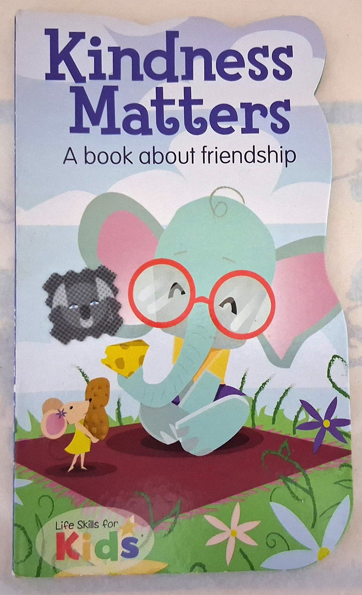 A children's book titled 'Kindness Matters: A book about friendship' with a colorful cartoon illustration of a light blue elephant wearing red glasses, sitting on a red blanket and sharing a yellow snack with a small mouse. The cover also shows the 'Life Skills for Kids' logo at the bottom left.