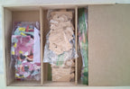 An open cardboard organizer box containing three compartments filled with craft or hobby pieces: a bag of colorful round pieces, a bag of small beige paper/cardboard pieces, and a stack of green-backed strips, with the right side showing the plain cardboard interior.