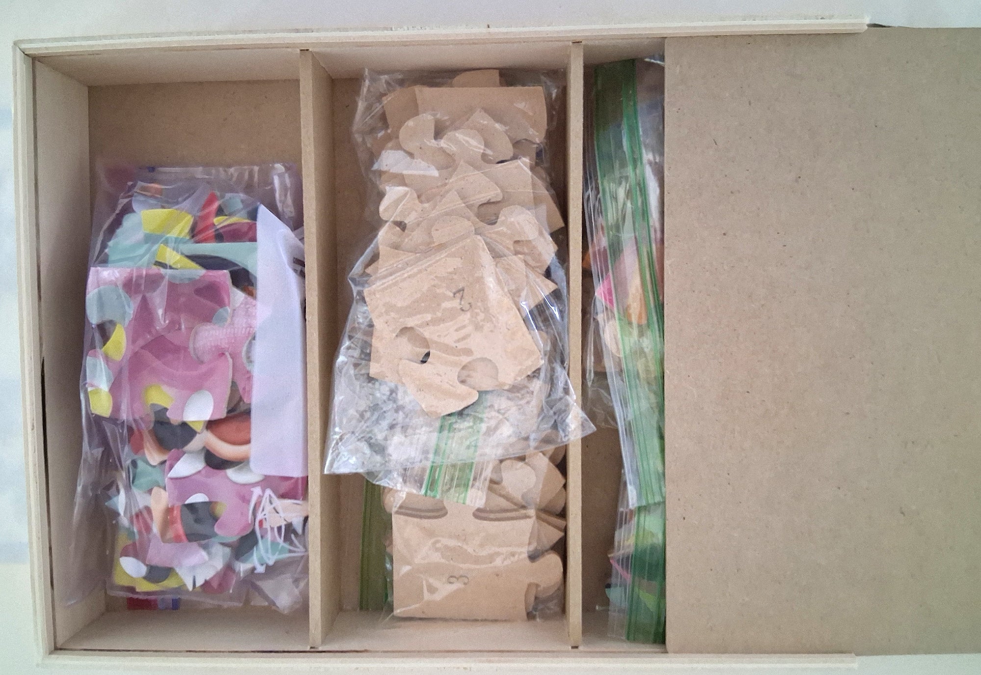 An open cardboard organizer box containing three compartments filled with craft or hobby pieces: a bag of colorful round pieces, a bag of small beige paper/cardboard pieces, and a stack of green-backed strips, with the right side showing the plain cardboard interior.