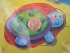 A colorful children's toy or craft featuring a cartoon turtle design, photographed close up on a yellow background. The turtle has a green shell with dark green spots, a pink center piece, blue legs and head, and a red outer shape.