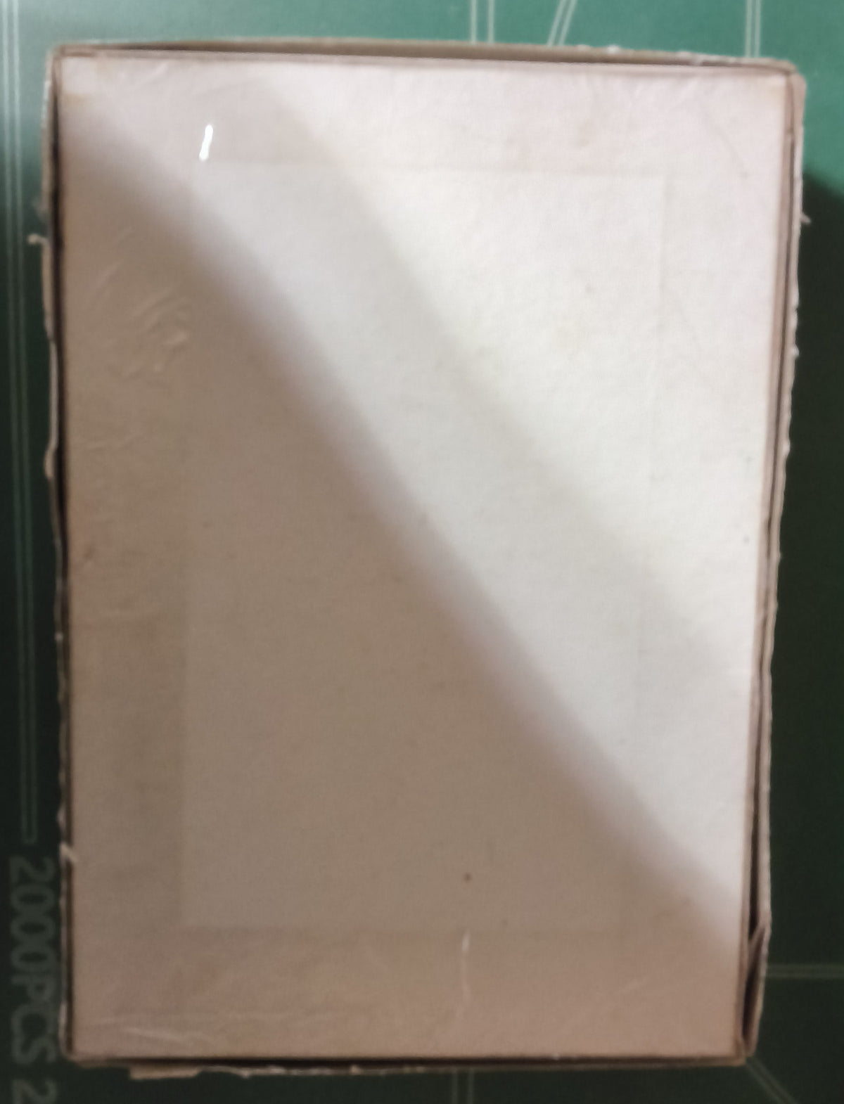 A plain white rectangular box or sheet centered on a green cutting mat with measurement markings and colorful letter stickers along the right edge.
